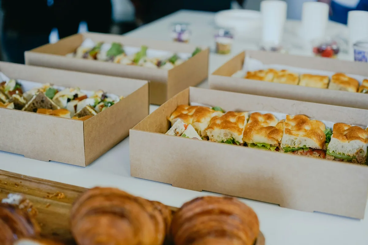 Kraft paper boxes of focaccia sandwiches and wraps set out on a white table, with croissants in the foreground.