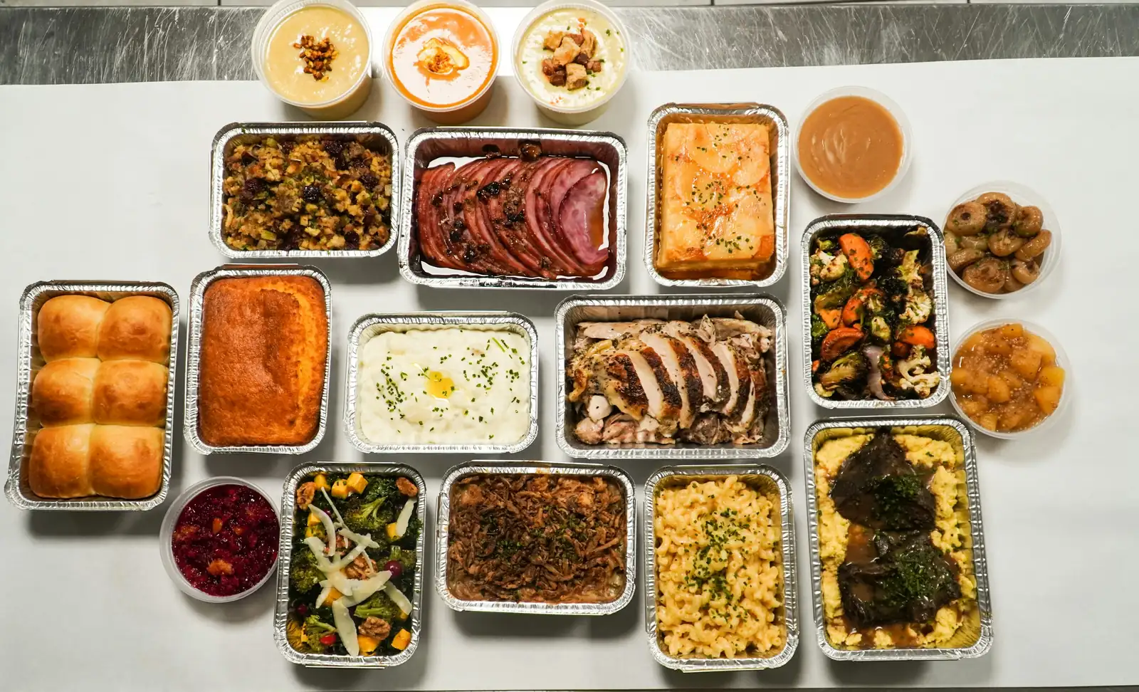 Overhead view of a full catering order in foil pans: sliced roast turkey, glazed ham, stuffing, mashed potatoes, mac and cheese, roasted vegetables, cornbread and dinner rolls, with soups and sides in round tubs.