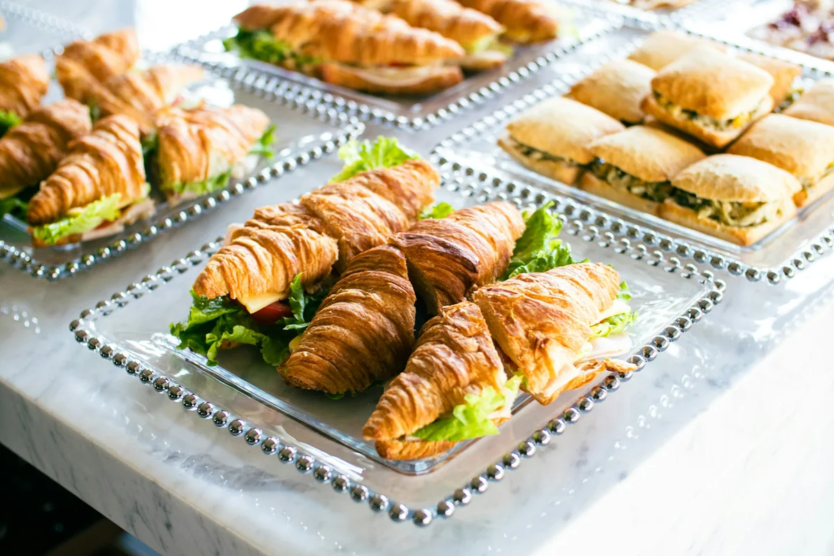 Glass platters of croissant sandwiches with lettuce and cheese beside a platter of filled sandwich rolls on a marble counter.