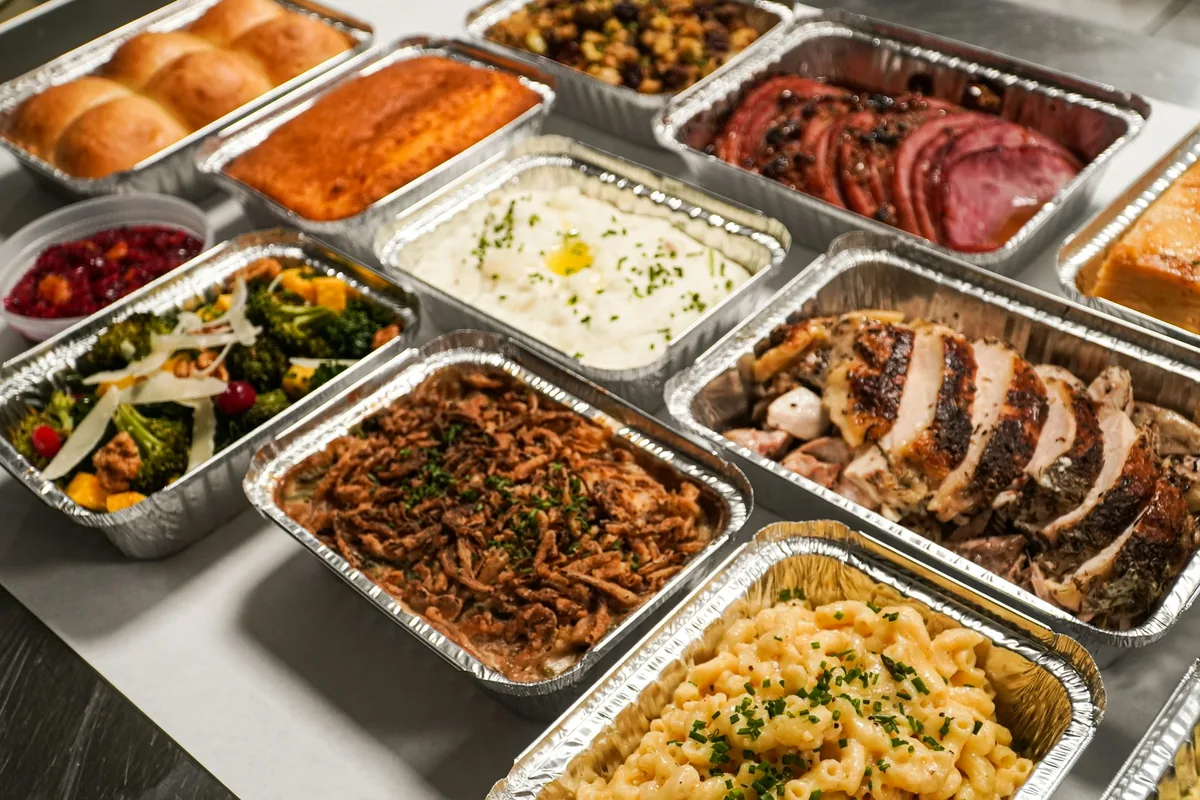 Foil pans of mac and cheese, green bean casserole, sliced roast turkey, mashed potatoes and dinner rolls lined up on a counter.