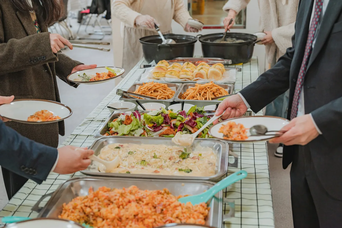 People in work clothes serve themselves from steel pans of seasoned rice, creamy pasta, salad, fries and rolls at a buffet line.