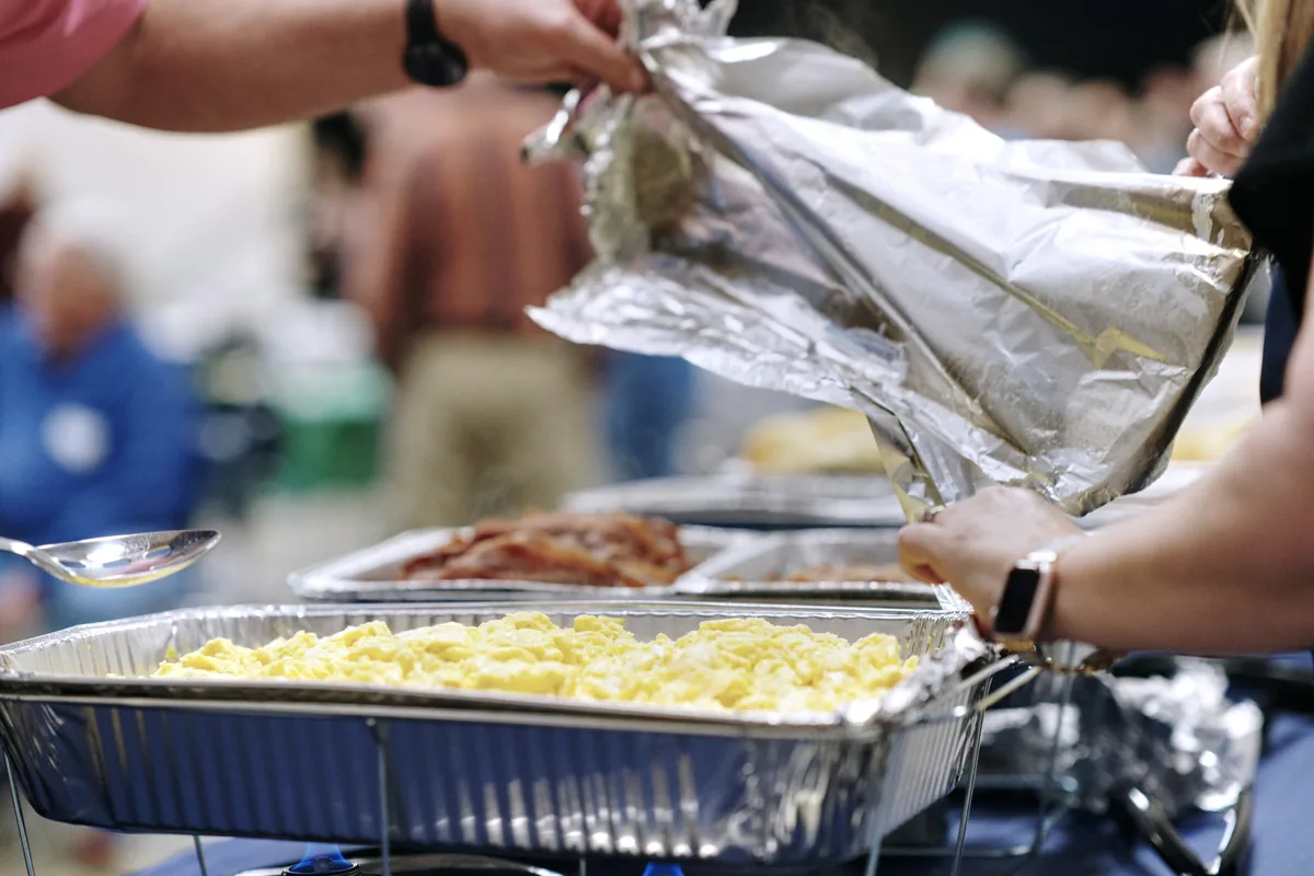 Hands peel the foil lid off a full pan of scrambled eggs in a wire chafing rack, with a pan of bacon behind it.